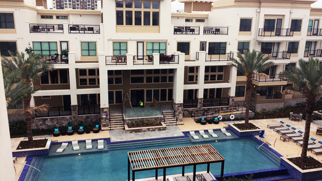 Modern apartment complex with a large outdoor swimming pool, surrounded by lounge chairs, palm trees, and balconies.